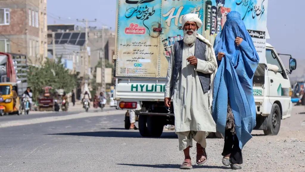 En afghansk man i traditionella kläder går bredvid en burkaklädd kvinna på en gata i utkanten av Herat. I bakgrunden syns en lastbil med färgglada reklamaffischer och stadsmiljö.