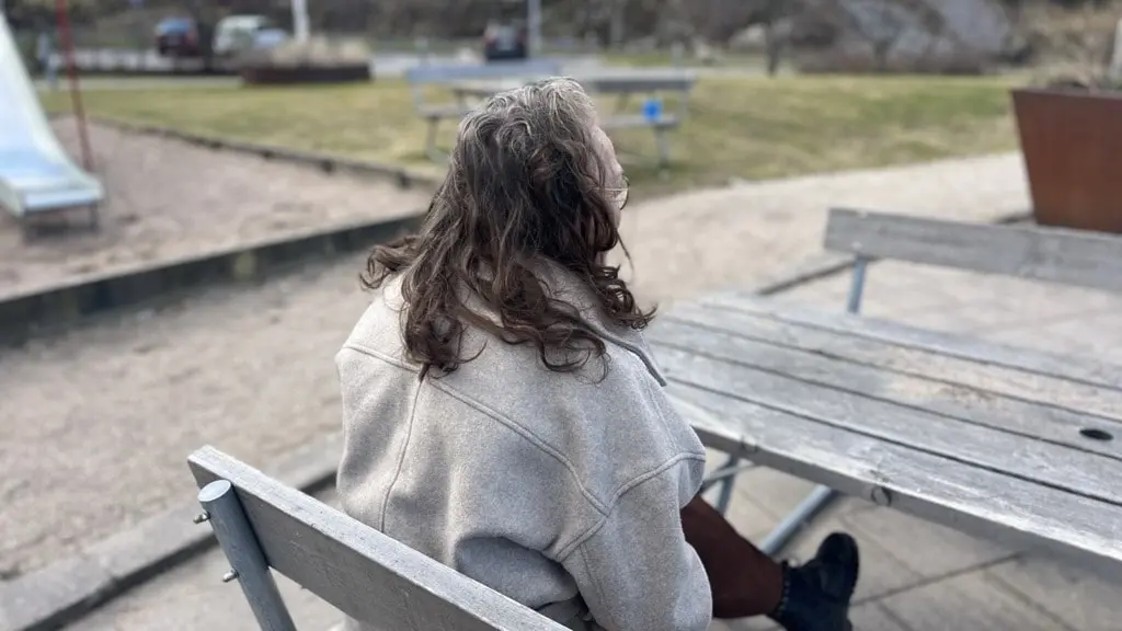 En kvinna med axellångt mörkt hår och glasögon sitter vid ett träbord utomhus i parkmiljö, fotograferad bakifrån i en ljusgrå jacka. 