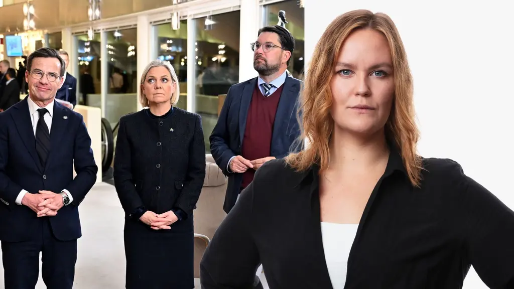 En grupp personer står inomhus, klädda i formella kläder. I förgrunden syns en person i svart kavaj mot en vit bakgrund. Bakgrunden har glasväggar och belysning.