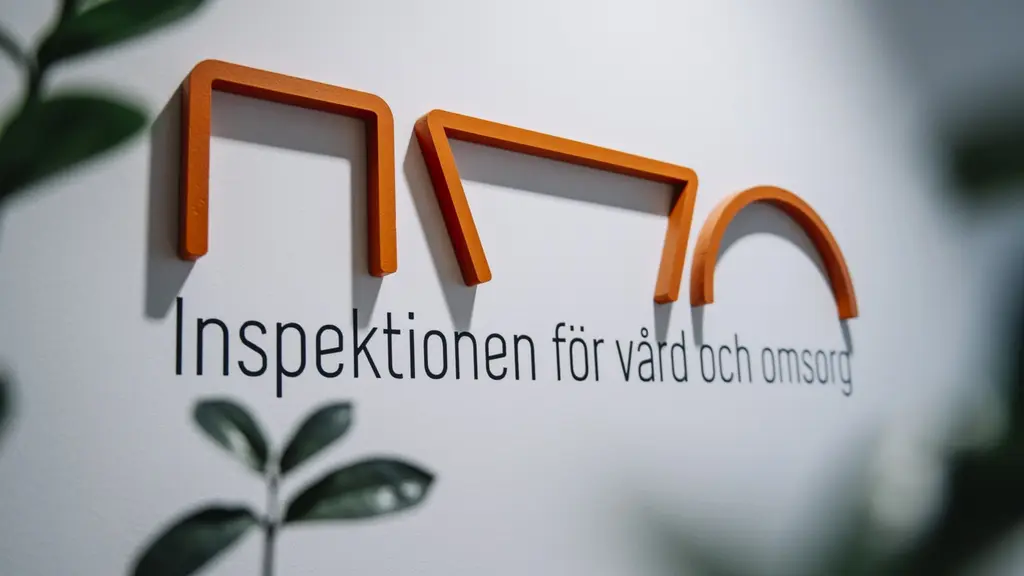 Logotypen för Inspektionen för vård och omsorg (Ivo) på en vit vägg. Logotypen är orange och består av geometriska former. I förgrunden syns gröna växtblad som skapar en naturlig känsla. 