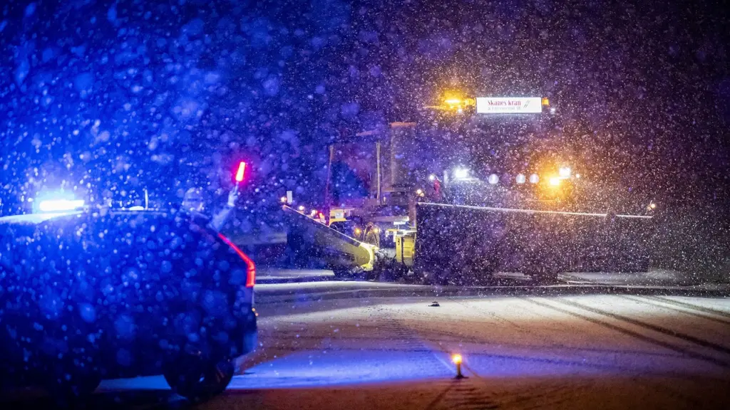 Kraftigt snöfall vid en trafikolycka på riksväg 110 nära Landskrona. En plogbil passerar och polisfordon med blåljus syns på plats, vilket lyser upp den vintermörka vägen.