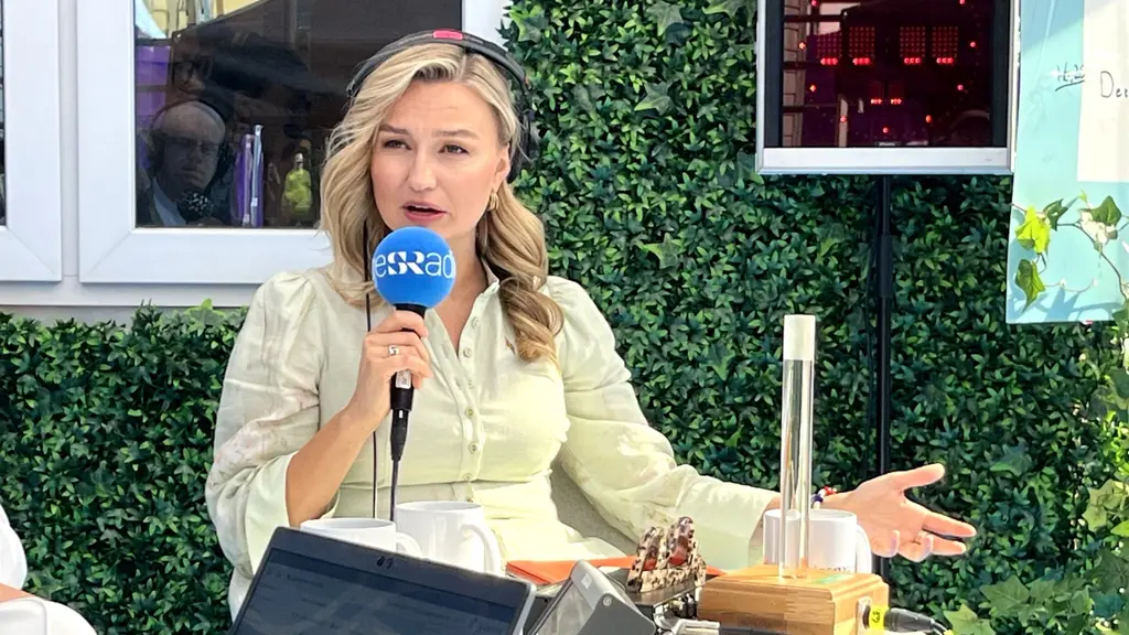 Kristdemokraternas partiledare Ebba Busch talar i mikrofon vid ett utomhusevenemang, sittandes vid ett bord och teknik framför en grön vägg med växtlighet.
