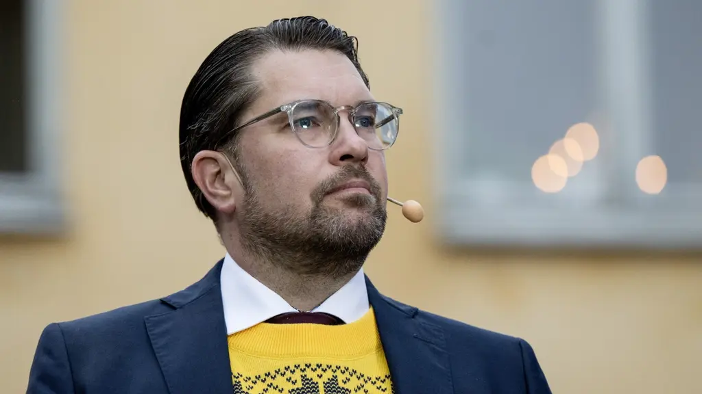 En person i blå kavaj och gul tröja talar via mikrofon under en pressträff utomhus vid en gul byggnad i Strängnäs. Bakgrunden visar ett suddigt fönster med ljuspunkter.
