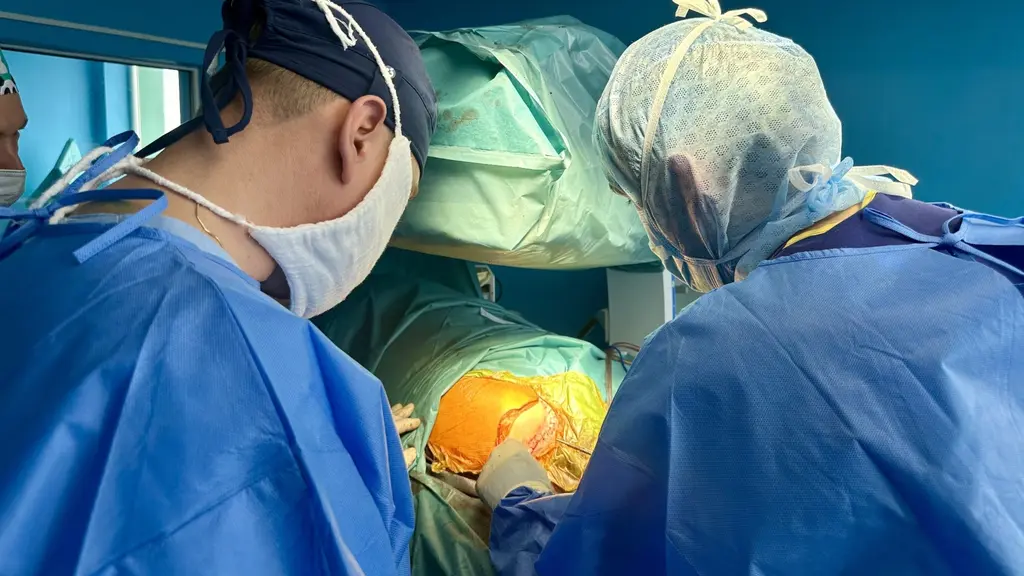 Kirurger utför en operation i en steril sjukhusmiljö. Två personer i blå kirurgkläder och hårskydd arbetar koncentrerat med en patient under stark belysning.