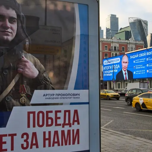 En affisch med texten 'Segern kommer att bli vår!' och en bild av en rysk soldat syns på en reklampelare. I bakgrunden kör bilar förbi en elektronisk billboard i en stadsmiljö med skyskrapor.