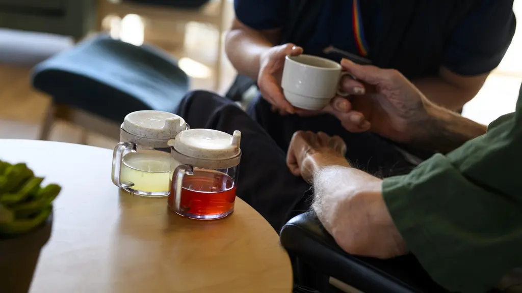 En äldre person får en kopp av en vårdare på ett äldreboende. I förgrunden syns pipmuggar och glasburkar med olika drycker på ett bord. Miljön är inomhus med mjuka möbler och trädetaljer.