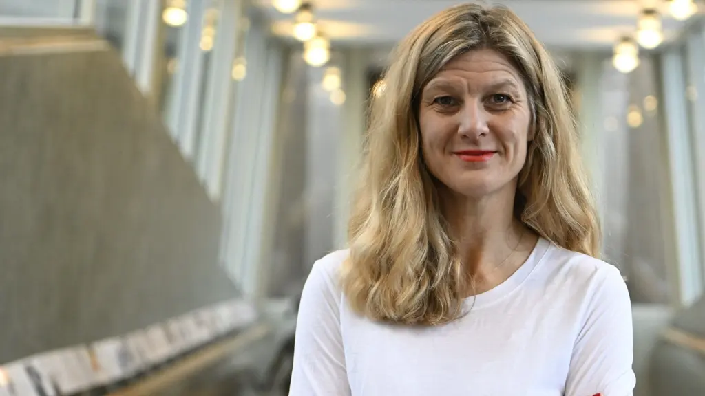 En kvinna med blont hår står i en ljus inomhusmiljö med glasväggar och långa gångar i bakgrunden. Hon är klädd i en vit tröja och har armarna i kors.