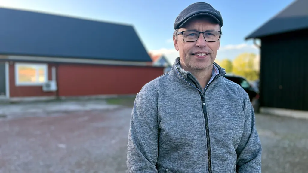 Man med glasögon i grå keps och tröja ler mot kameran med en röd ladugårdsliknande byggnad bakom sig.