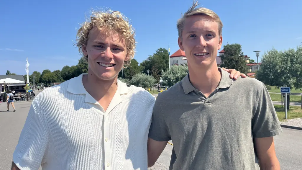 Två personer står bredvid varandra utomhus på en solig dag. Bakgrunden visar en grönskande park, en flagga och byggnader. De är klädda i ljusa, somriga kläder.