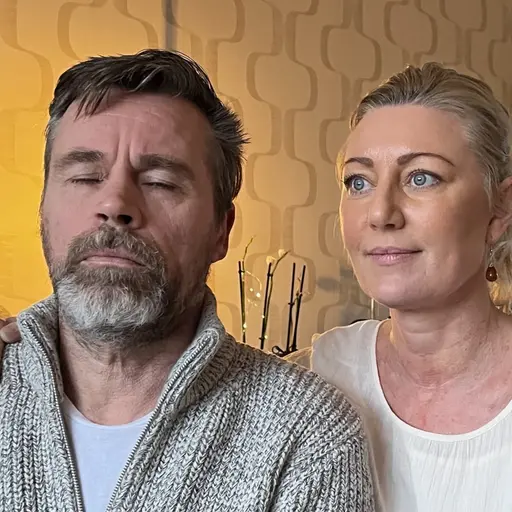 En man och en kvinna sitter tillsammans i ett vardagsrum. Mannen blundar och kvinnan tittar bort mot ljuset från ett fönster. Hon håller sin ena arm kring mannen. Han har kort hår och ett silvrigt, ganska kort skägg. Hon har blont uppsatt hår. Dom har ljusa kläder.