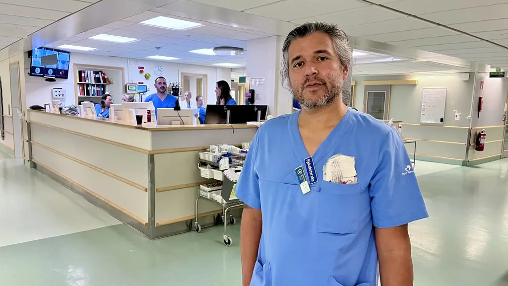 Eric Åkerman står i sjukhusmiljö iklädd blå arbetskläder. Bakgrunden visar reception och personal i arbete.