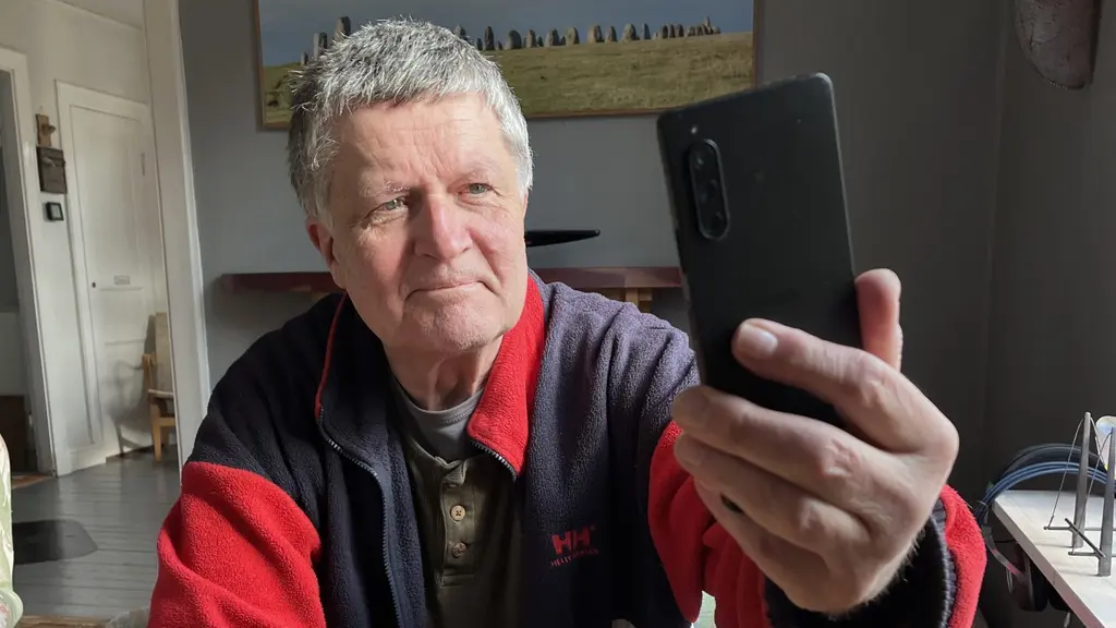 Mikael Johansson sitter och håller upp sin mobiltelefon framför sig i ett rum.