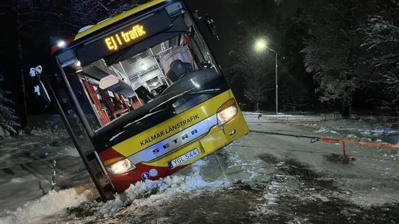 En gul KLT-buss med texten 'Ej i trafik' har kört av vägen i Emmaboda och lutar kraftigt mot en snöig vägkant. En vajer är fäst vid bussen för att försöka dra upp den.