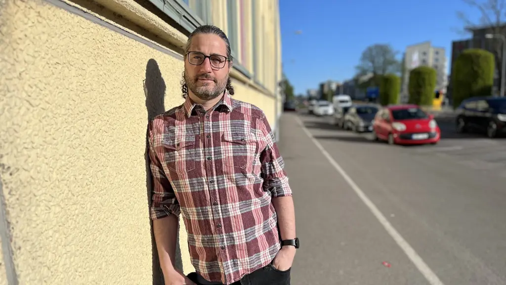 Porträtt på man som står lutad mot en gul putsfasad. Det starka solljuset får kroppen att skapa en skugga mot fasaden. I bakgrunden kör bilar på den intilliggande bilvägen. Han har håret uppsatt i en knut och glasögon.
