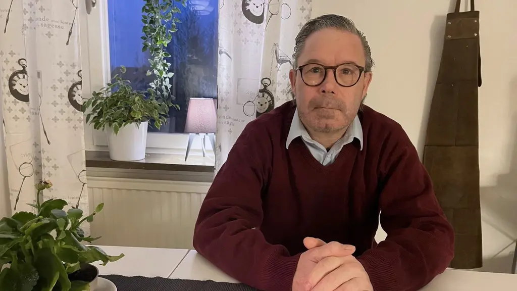 En man i mörkröd tröja sitter vid ett bord i ett kök. Väpnat rån trasade sönder Marcus liv: ”Man blir iskall”