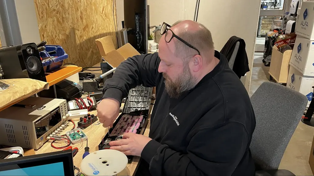 En person sitter vid ett arbetsbord fyllt med elektronisk utrustning och arbetar med att montera eller reparera en teknisk komponent. Rummet innehåller elektronisk utrustning och verktyg.