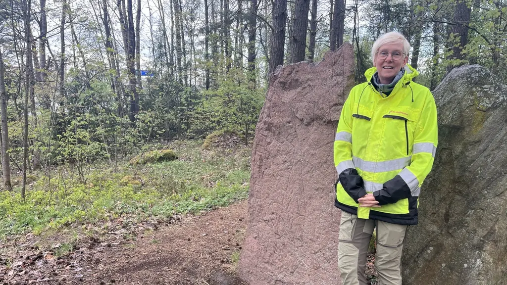 En kvinna i reflexjacka står vid en runsten. Åsa berger vid runstenen. Arkeologisk utgrävning av gamla gravar vid rondellen