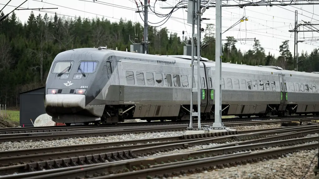 Ett snabbtåg åker på en järnväg. I bakgrunden syns skog och i förgrunden elledningar.