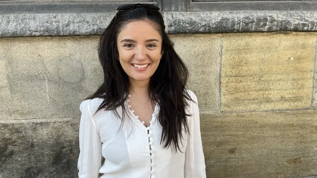 Sussan Rahmani står framför en stenvägg klädd i vit blus. Hon har långt mörkt hår och solglasögon på huvudet.