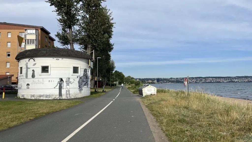 Vy över Vätterstranden med en gång- och cykelväg som sträcker sig längs vattnet. En vit byggnad med muralmålning syns på vänster sida och sjön Vättern ligger till höger.