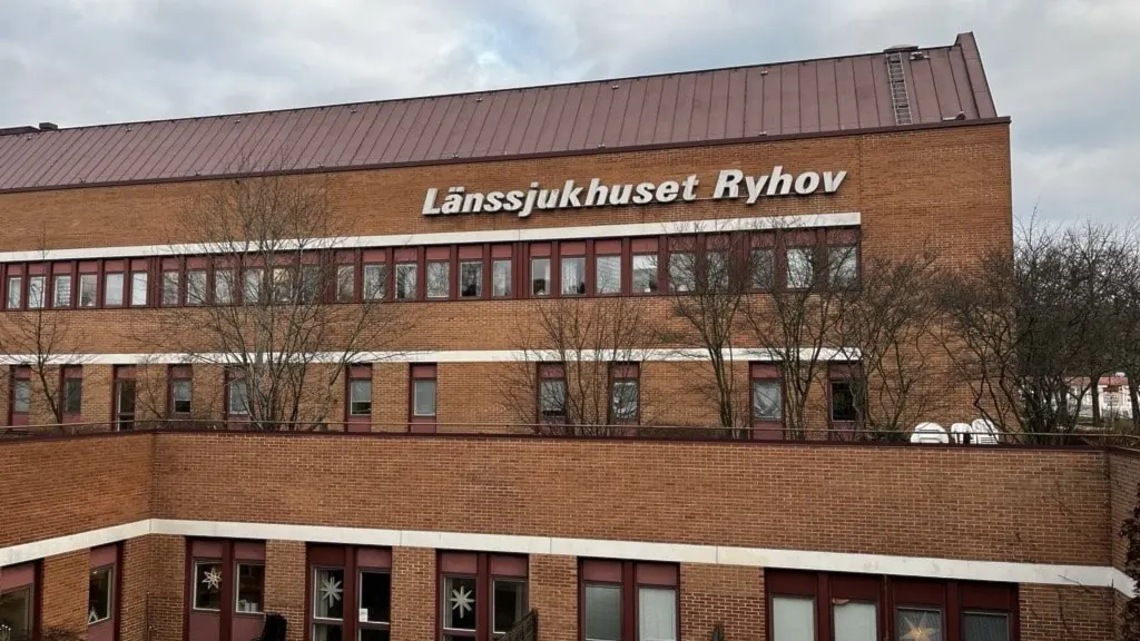Bild på Länssjukhuset Ryhov, en brun tegelbyggnad med texten 'Länssjukhuset Ryhov' på fasaden. Framför byggnaden syns träd och buskar. Himlen är molnig i bakgrunden.