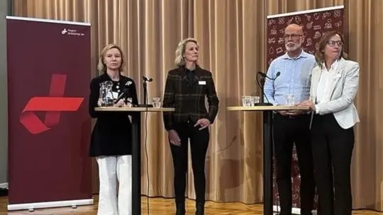 Fyra personer står på en scen med mikrofoner och podium under en paneldiskussion. Bakom dem syns två rollups och beigea gardiner. Diskussionen verkar vara kopplad till ett event eller konferens.
