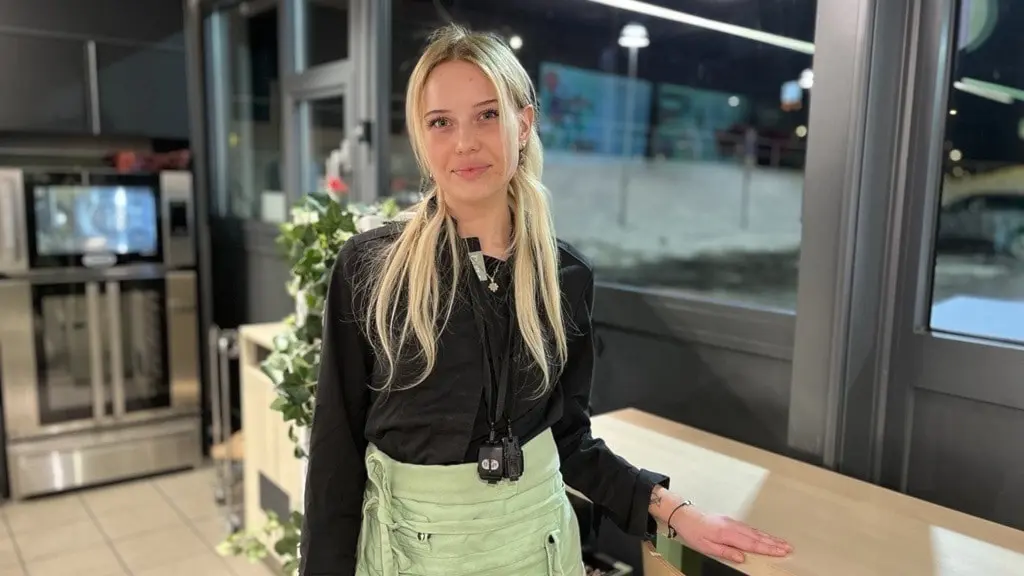 En kvinna står inomhus i ett rum med stora fönster som visar att det är mörkt utomhus. Hon bär en ljusgrön förkläde över mörka kläder och håller ut ena handen mot ett bord.