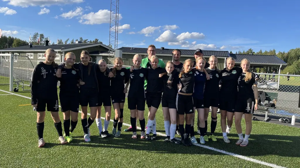 Ett fotbollslag från Forserums IF poserar på en gräsplan under en klar blå himmel. Laget består av unga spelare i svarta träningskläder, med en byggnad och fotbollsmål i bakgrunden.