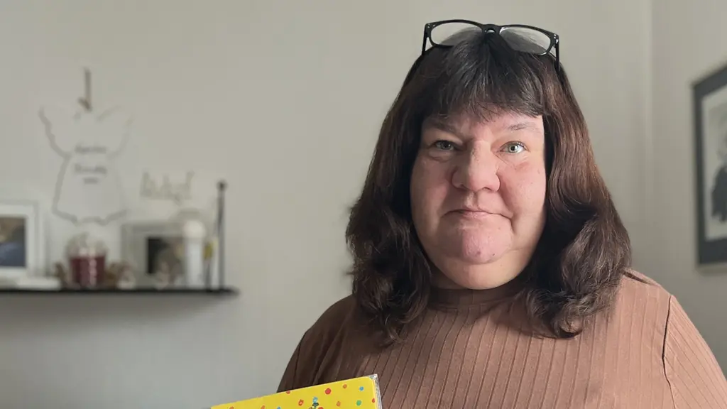 En person iklädd brun tröja håller en barnbok i ena handen och en rosa bricka med leksaksmat i den andra. Bakgrunden visar en heminredning med ljusa väggar och en gardin.