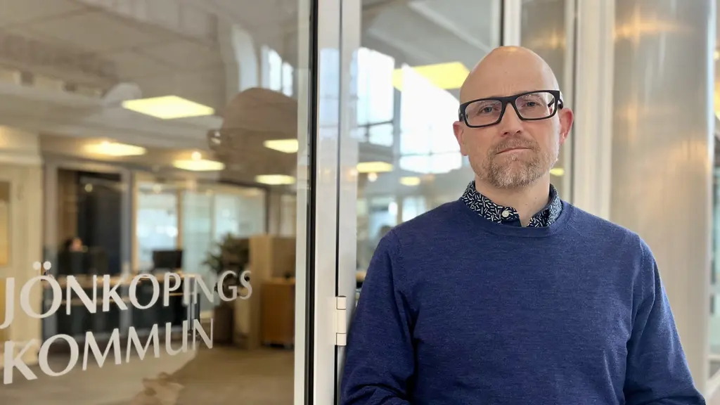 En person står framför en glasdörr med texten 'Jönköpings kommun'. I bakgrunden syns ett kontorslandskap med skrivbord och belysning.