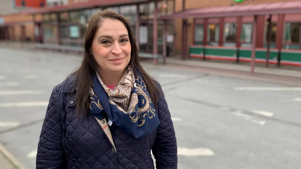 Rachel de Basso står utomhus vid en gata med färgglada byggnader i bakgrunden. Hon bär en mörkblå jacka och en mönstrad scarf.