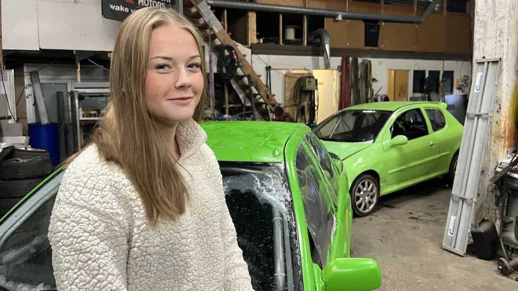 En bilverkstad med två gröna bilar i fokus. En person med långt hår står framför en av bilarna. Lokalen är fylld med verktyg och utrustning, samt hyllor med förvaringsmaterial.
