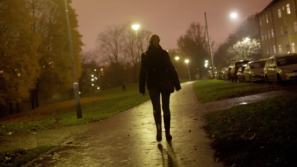 En kvinna promenerar ensam på en regnvåt gångväg iunder en mörk kväll. Gatlyktor lyser upp omgivningen med ett varmt sken medan träd och bostadshus skymtar i bakgrunden.