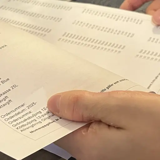 En person håller i ett papper med en lista, medan fler dokument ligger på bordet i bakgrunden. Miljön verkar vara inomhus och fokus är på händerna och dokumenten.