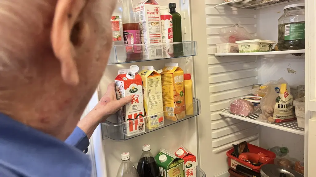 En äldre man, i 85-årsåldern, står vid ett kylskåp och tittar på en hylla fylld med mjölkförpackningar, juice och andra livsmedel. Kylskåpet innehåller olika matvaror, inklusive drycker och burkar.