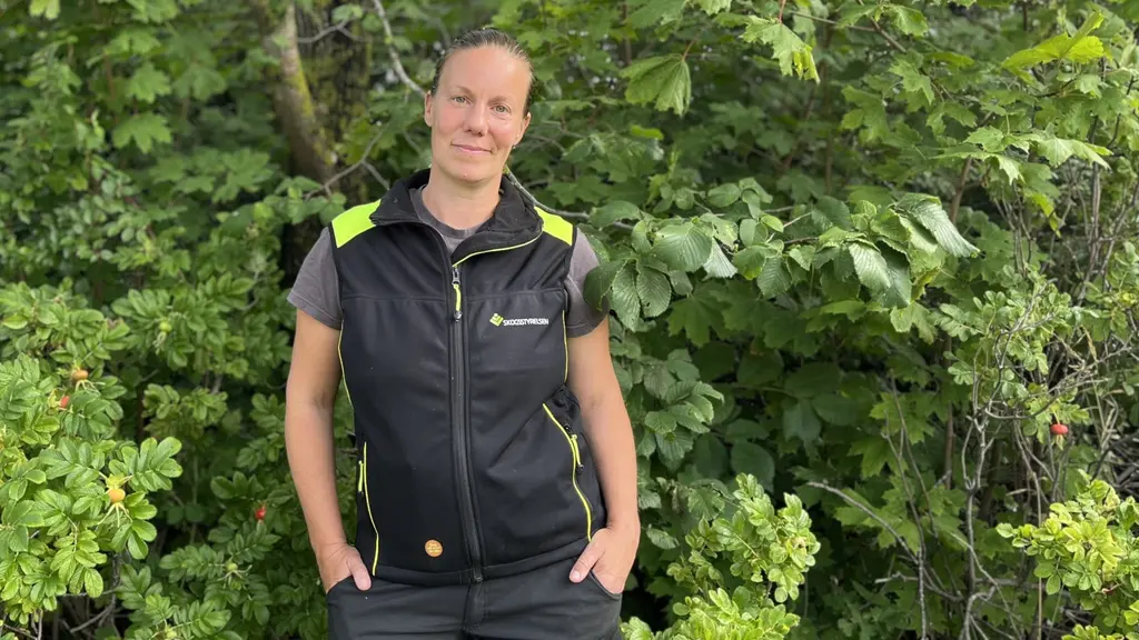 Amanda Overmark står utomhus framför grönskande buskar och träd, iklädd en mörk arbetsväst med gula detaljer. Hon har händerna i fickorna och omgivningen är frodig och somrig.