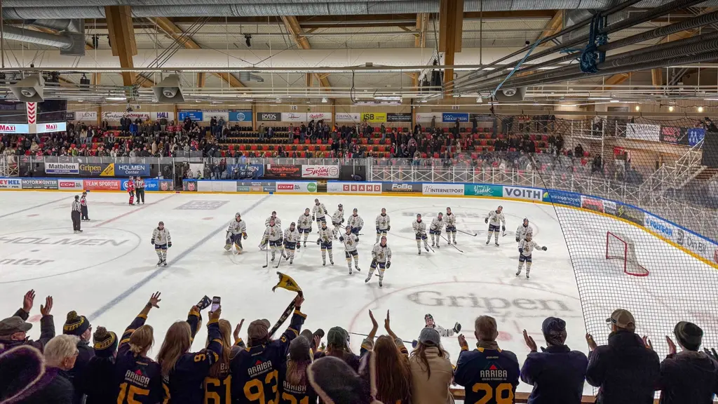 Ishockeyspelare står på isen och tackar publiken efter en match. Arenan är fylld med åskådare, och supportrar med lagtröjor syns i förgrunden. Banderoller och reklam syns längs sargen.