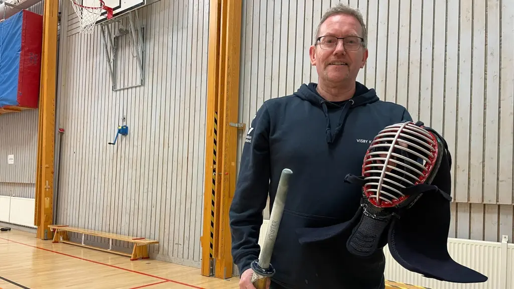 En person står i en idrottshall och håller i en kendohjälm och ett träningssvärd. Bakgrunden visar ribbväggar, en basketkorg och träningsmattor. Personen är klädd i mörka kläder.