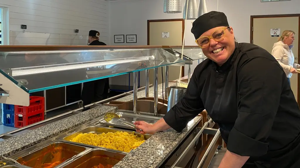 Martina Jakobsson, krigsplacerad storkökskock, står vid en buffédisk med flera rätter. I bakgrunden syns en ljus restaurangmiljö. 