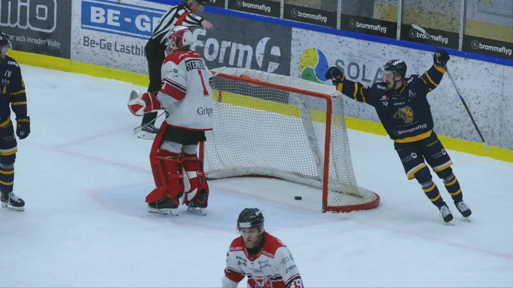 En ishockeyspelare firar ett mål med utsträckta armar medan motståndarlagets målvakt står framför målet. En annan spelare från motståndarlaget syns nära målet. Matchen äger rum på en inomhusrink.