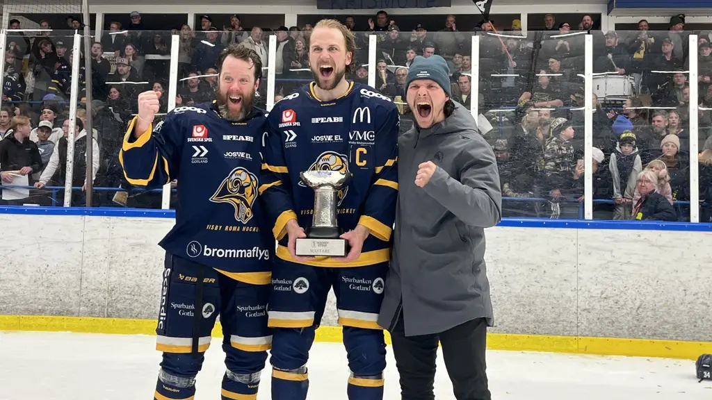Tre personer står på en ishockeyrink, varav två är klädda i blåa hockeyuniformer med gul detaljer och den tredje i en grå jacka. De ser glada ut och håller en pokal. Publik syns i bakgrunden.