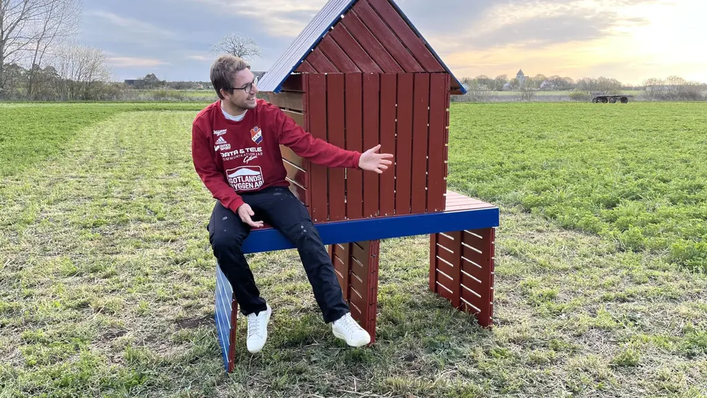 En man sitter på en del av ett rött miniatyrhus, med två hål under. Han pekar med armen mot resten av huset. Huset står på ett öppet, grönt fält. 