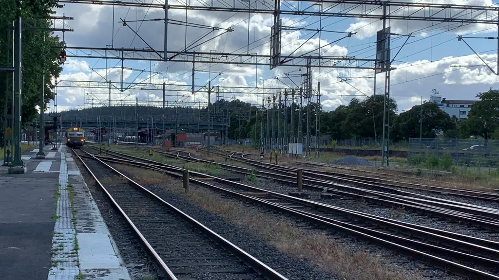 Den långa perrongen på spår 1 sträcker sig brevid järnvägspåret, längre bort står ett tåg. Flera järnvägspår med växlar mellan. Mot himlen finns flera kotaktledningar och elstolpar i stål. Den blå himlen döljs mestadels av moln. 