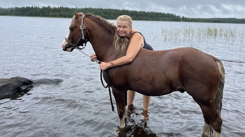 Maria Ohlsson står i vattnet med en brun häst vid en sjö. Bakgrunden visar ett skogsområde och gråa moln på himlen, vilket skapar en naturlig och stillsam miljö.