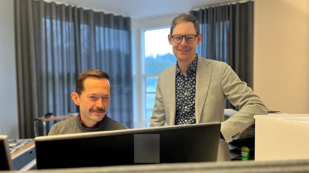 Två personer står vid ett skrivbord med datorer i ett kontor. Bakgrunden visar stora fönster med grå gardiner som släpper in dagsljus.