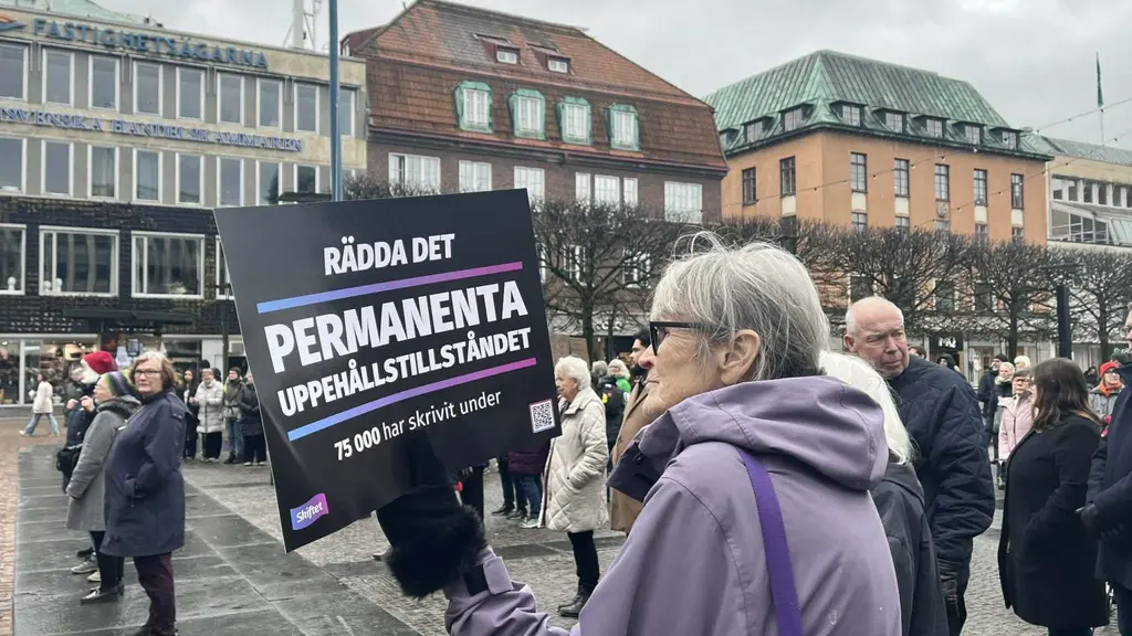 En demonstration på ett torg med en kvinna som håller en skylt med texten 'Rädda det permanenta uppehållstillståndet'. Bakgrunden visar byggnader och en samling människor.