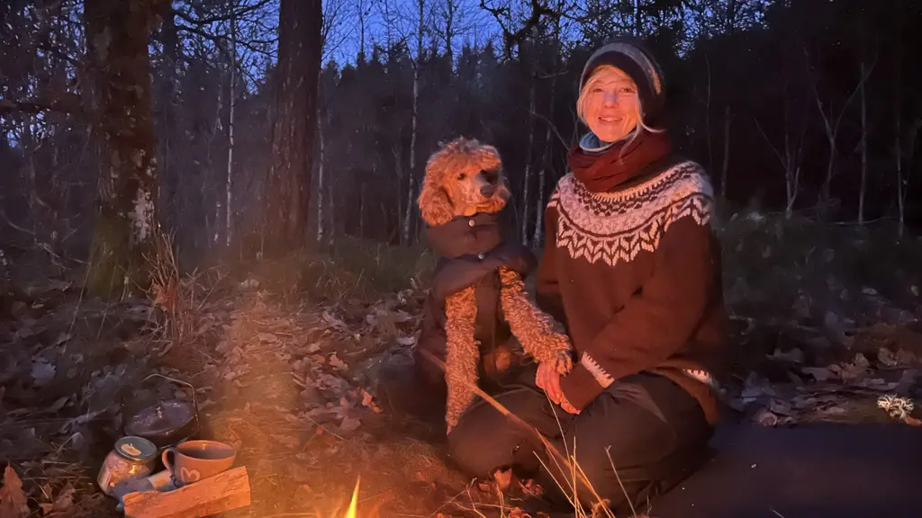 En person i varm stickad tröja sitter utomhus med en liten hund iklädd täcke. De är omgivna av skog och löv, och en eld brinner framför dem.