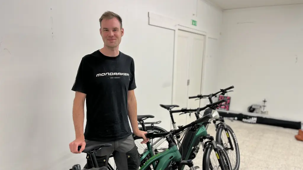 Edvin Wilson står i ett rum med flera elcyklar, klädd i en svart t-shirt och shorts. Han håller i en av cyklarna som är grön och svart.