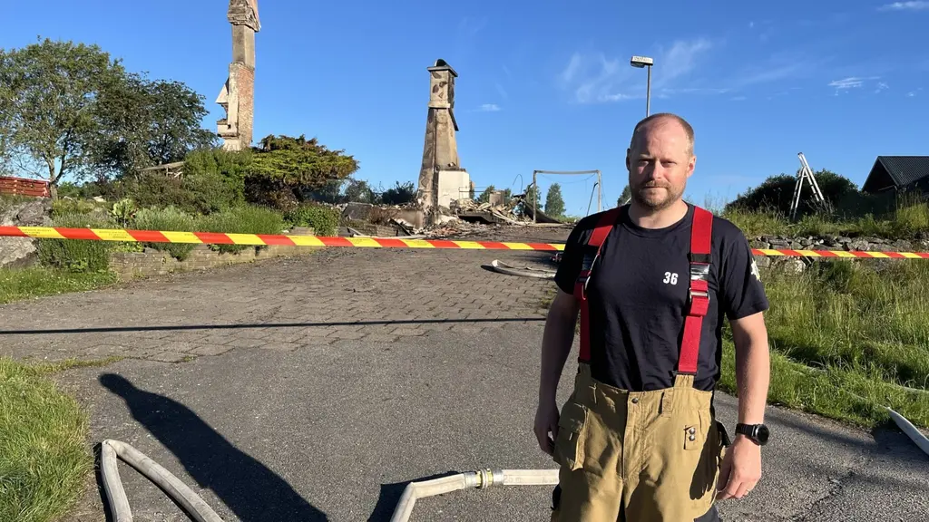 Brandman står vid avspärrning framför rasmassor och två skadade skorstenar efter kraftig brand, i ett villaområde under klar himmel.