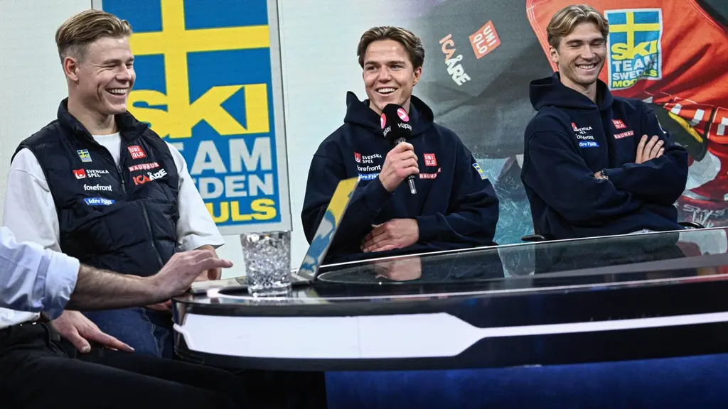 Tre puckelpiståkare sitter vid ett podium klädda i svenska landslagets kläder under Svenska Skidförbundets medieupptakt i Stockholm. I bakgrunden syns en svensk flagga och skidförbundets logotyp.